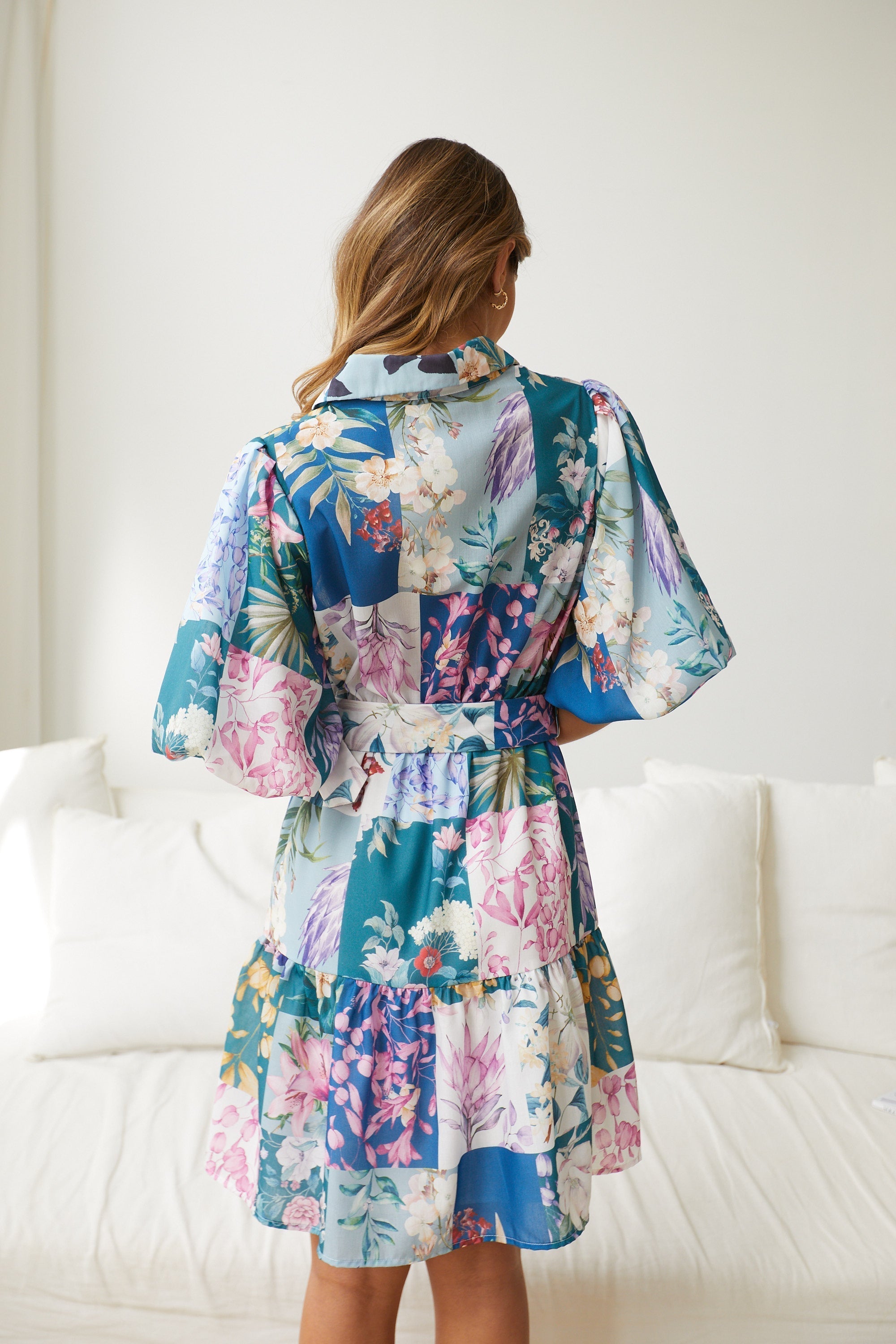 A woman wearing a short, multicolored floral dress with puff sleeves and a belted waist. The dress features a patchwork design with vibrant flower patterns in shades of pink, blue, yellow, and green. She is standing indoors in front of a white couch, looking to the side with a gentle smile.