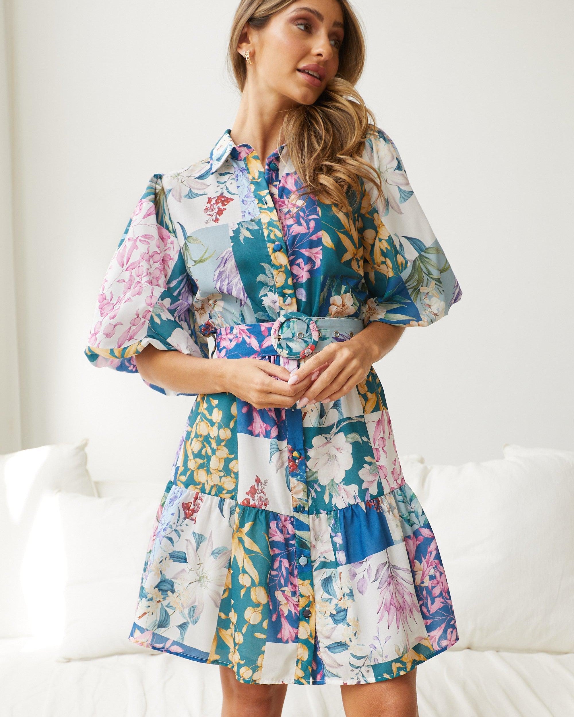 A woman wearing a short, multicolored floral dress with puff sleeves and a belted waist. The dress features a patchwork design with vibrant flower patterns in shades of pink, blue, yellow, and green. She is standing indoors in front of a white couch, looking to the side with a gentle smile.