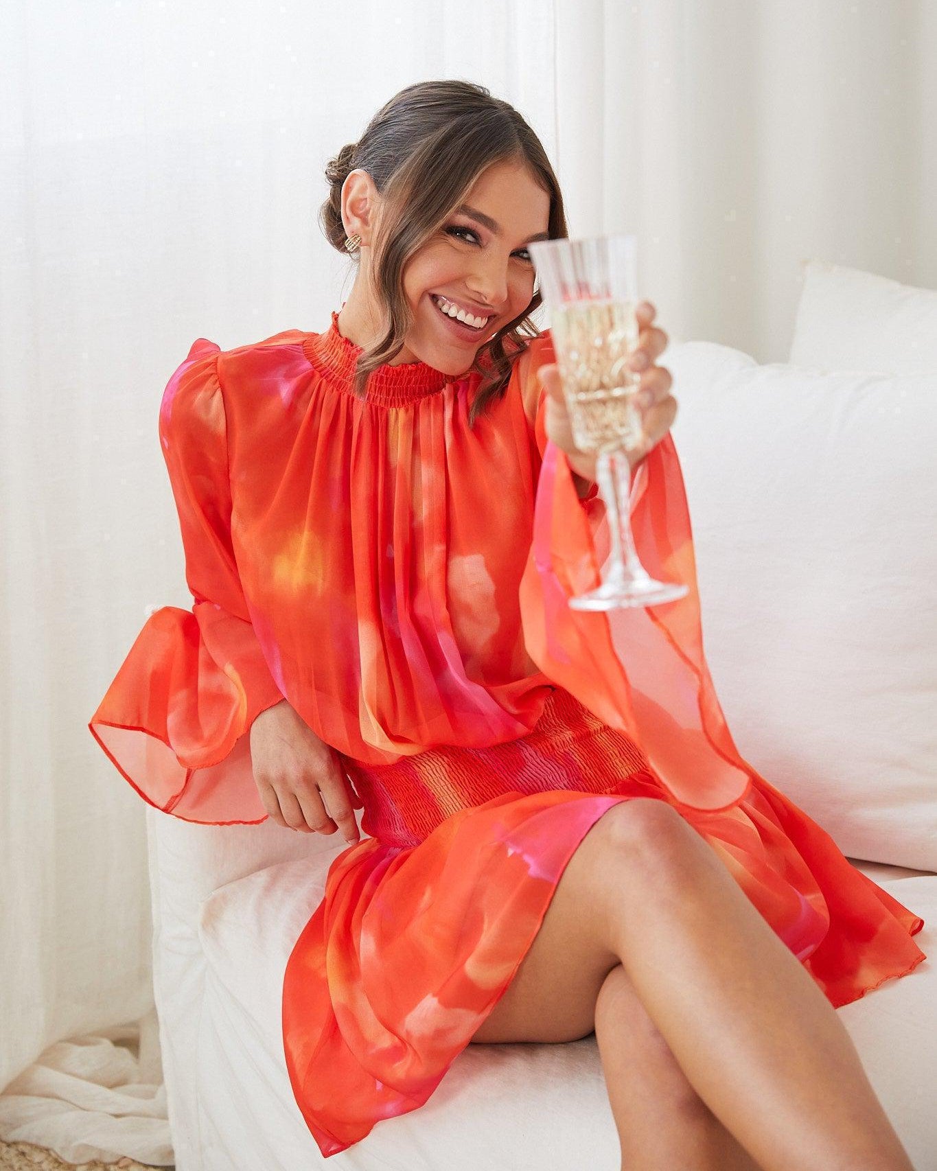 A woman sitting on a white couch, holding a glass of champagne, wearing an orange base dress with a red and purple print, featuring a high neckline and long trumpet sleeves.