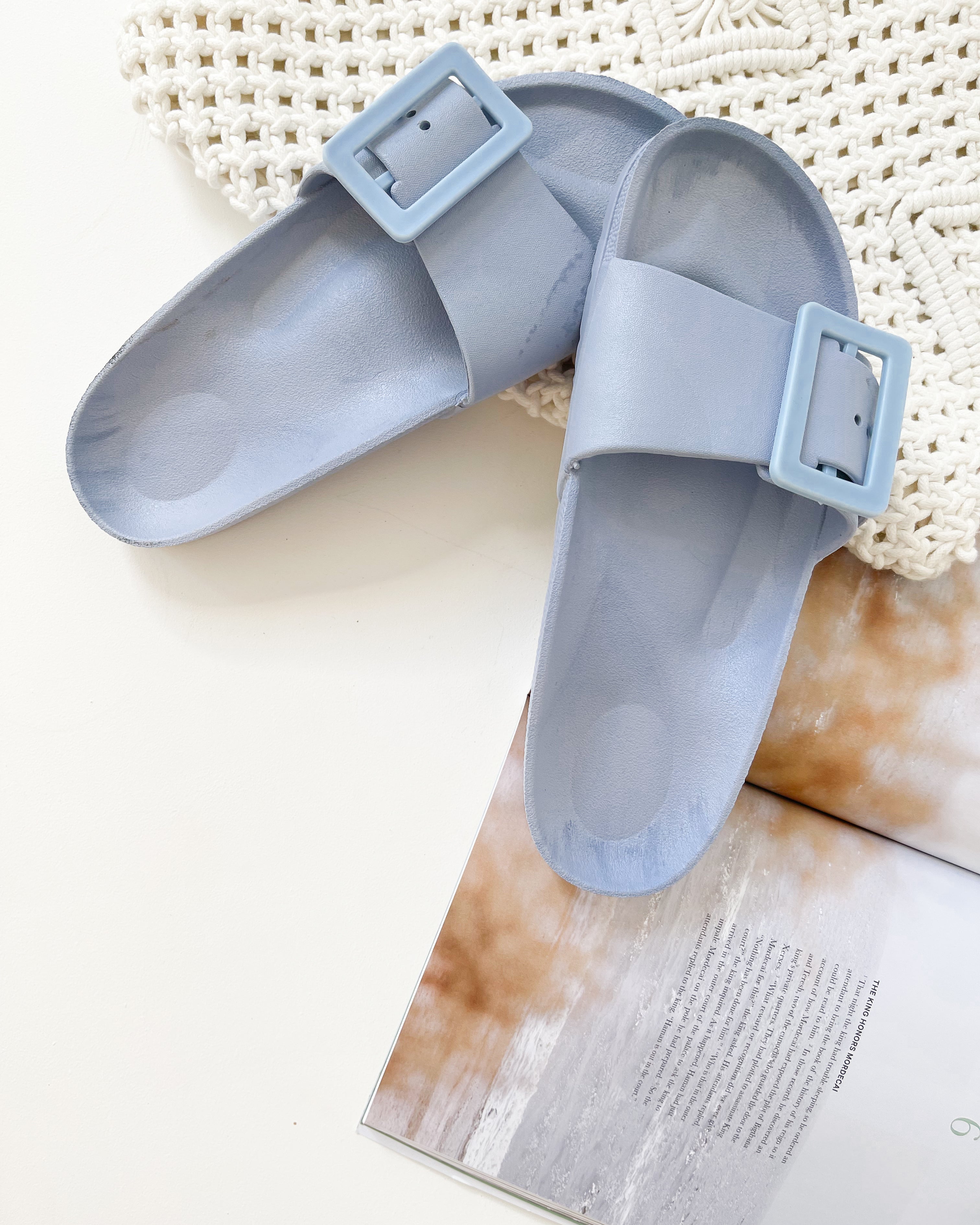 A pair of black summer slides with thick straps, displayed on a flat surface with a knitted texture in the background.