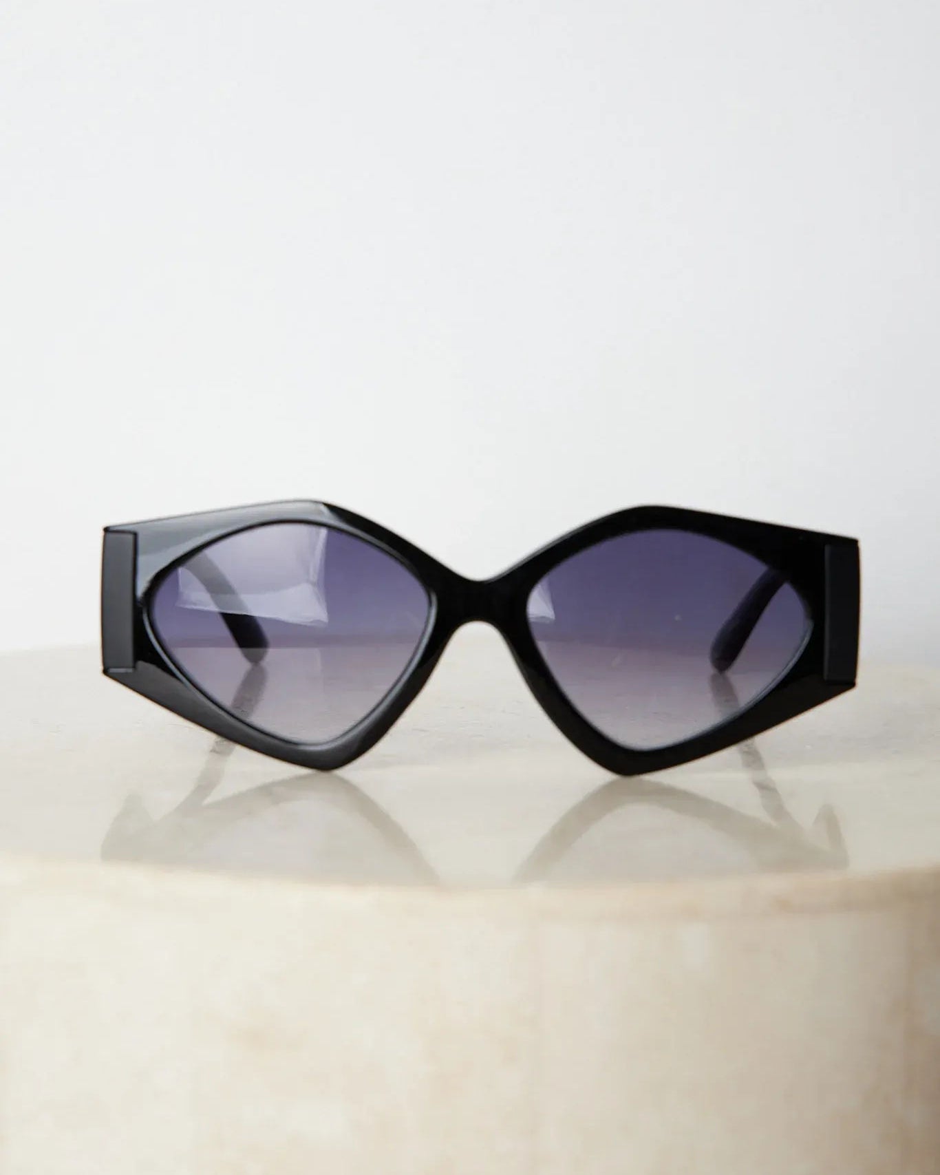 Black cat-eye sunglasses with thick frames and dark tinted lenses, displayed on a light beige surface against a plain white background.
