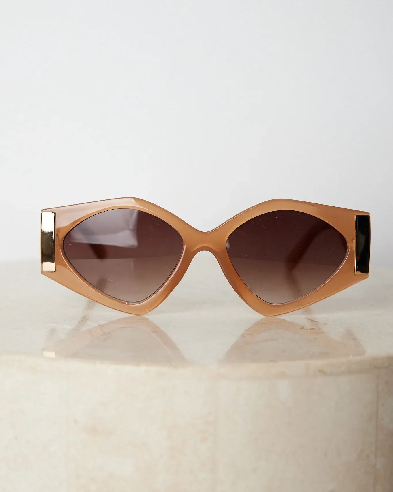 Brown cat-eye sunglasses with thick, glossy frames and dark brown tinted lenses, featuring gold accents on the corners, displayed on a light beige surface against a plain white background.