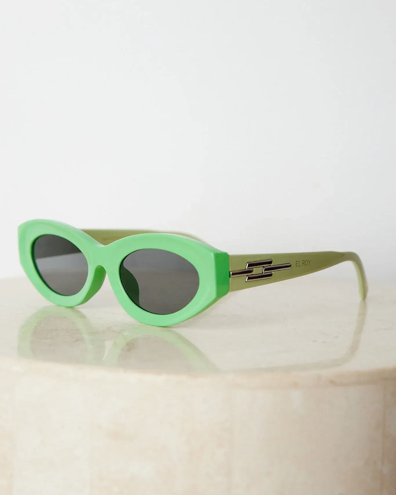 Close-up of a pair of bright green cat-eye sunglasses with dark lenses, placed on a smooth beige surface with a plain white background.