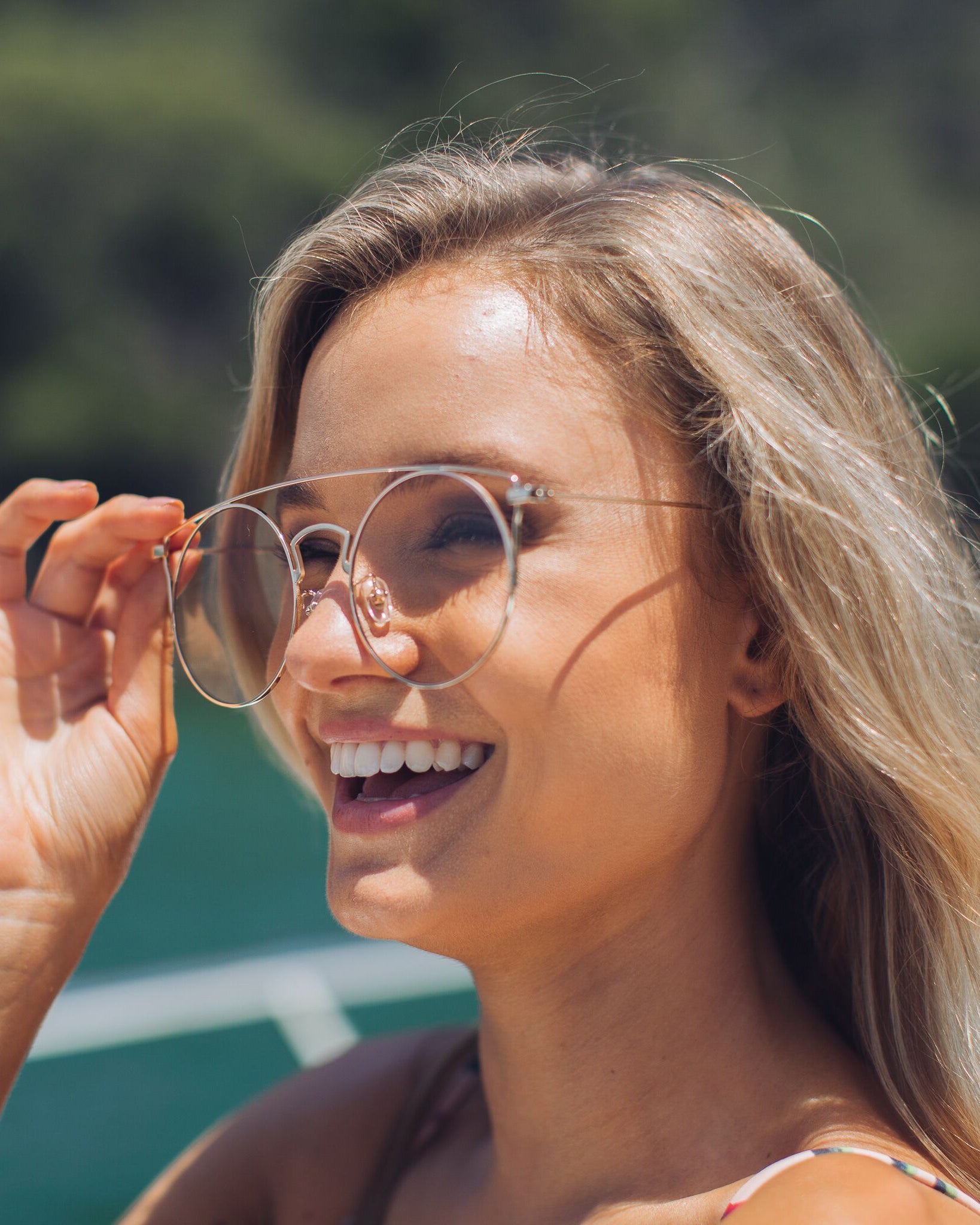 Close-up of a hand holding a pair of gold-framed round eyeglasses with a double bridge and black temple tips. The background shows a light wooden floor and a woman’s legs in white shorts and beige open-toe heels, slightly out of focus.