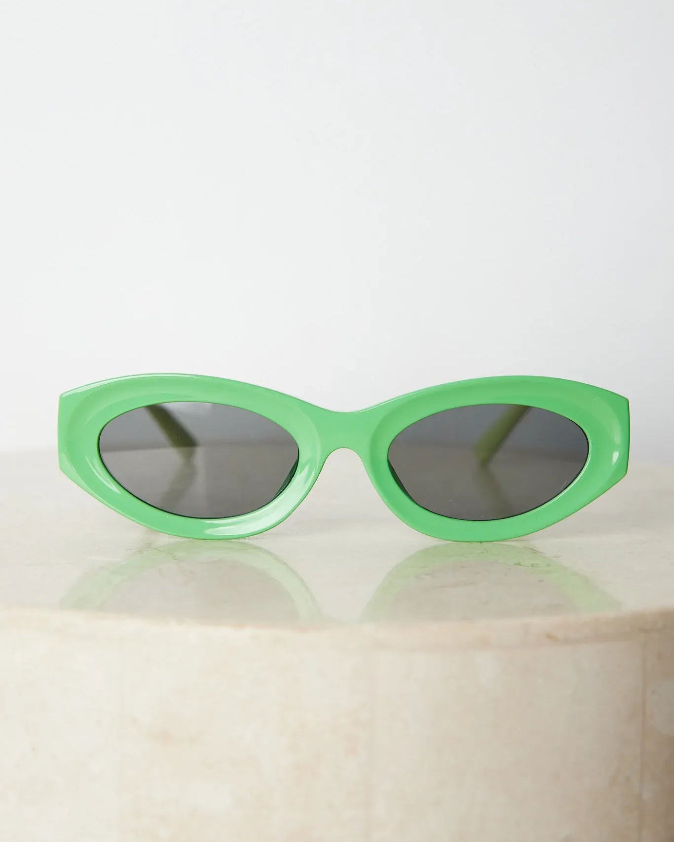 Close-up of a pair of bright green cat-eye sunglasses with dark lenses, placed on a smooth beige surface with a plain white background.