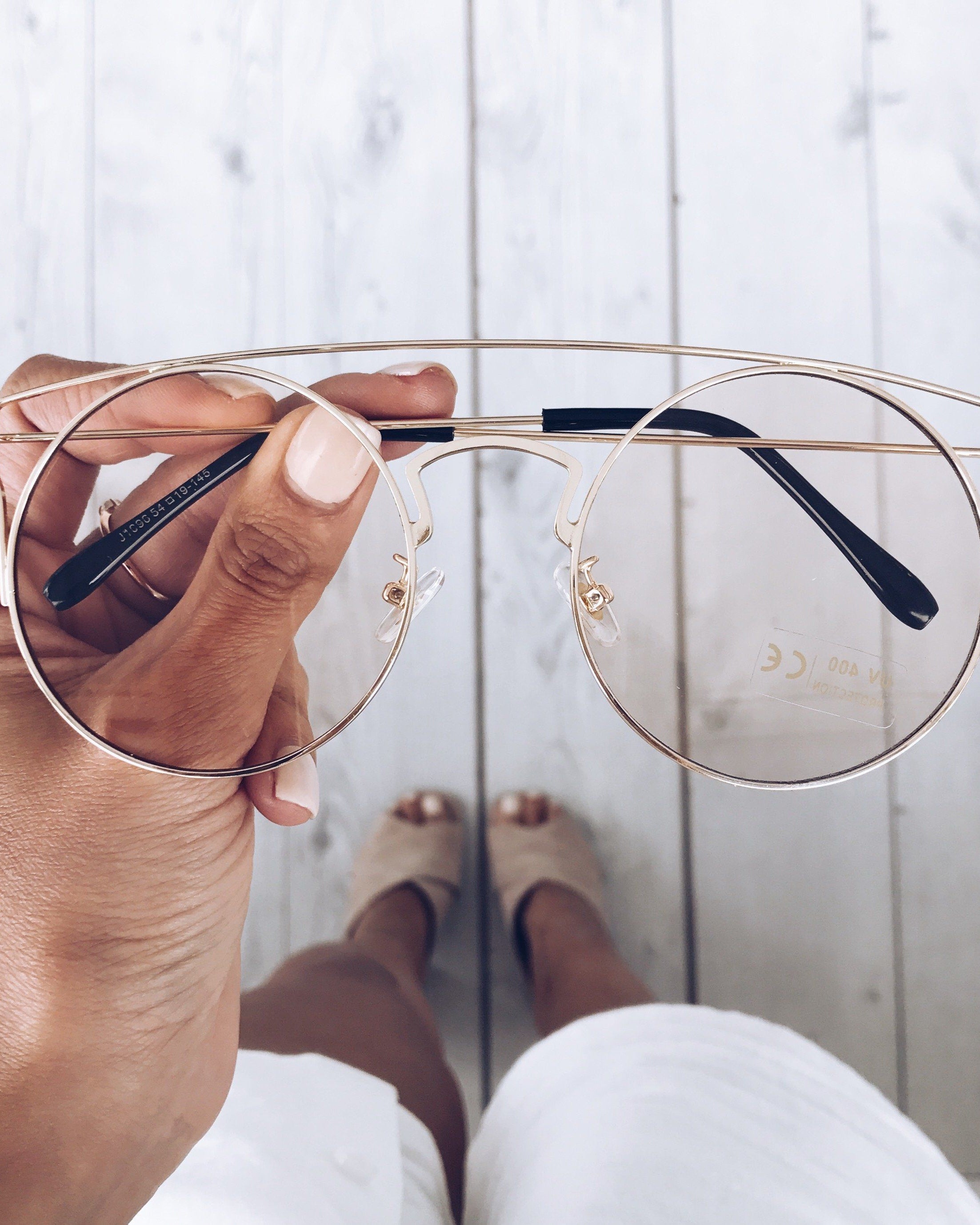 Close-up of a hand holding a pair of gold-framed round eyeglasses with a double bridge and black temple tips. The background shows a light wooden floor and a woman’s legs in white shorts and beige open-toe heels, slightly out of focus.