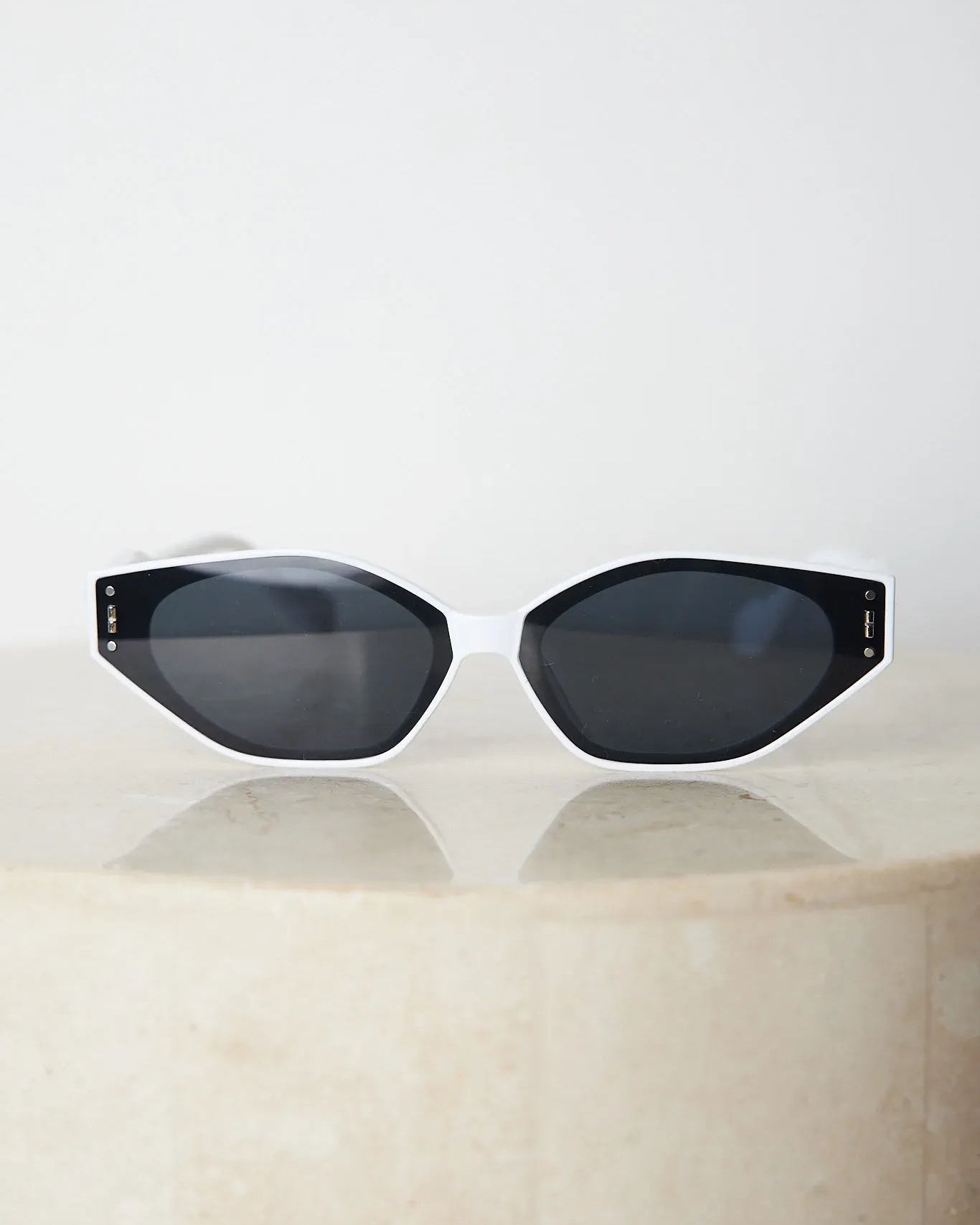 White geometric-shaped sunglasses with dark lenses resting on a light marble surface against a plain white background.