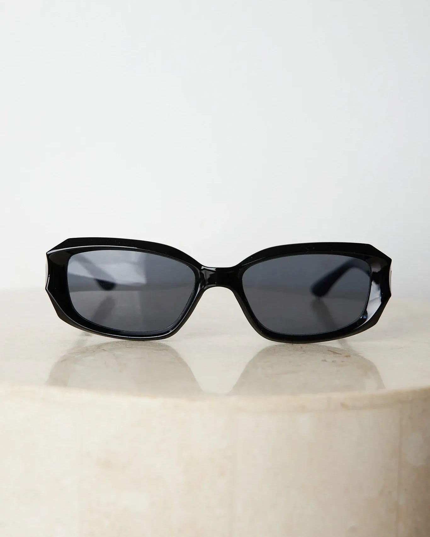 Black rectangular sunglasses with a glossy frame and dark tinted lenses, displayed on a light beige curved surface against a plain white background.
