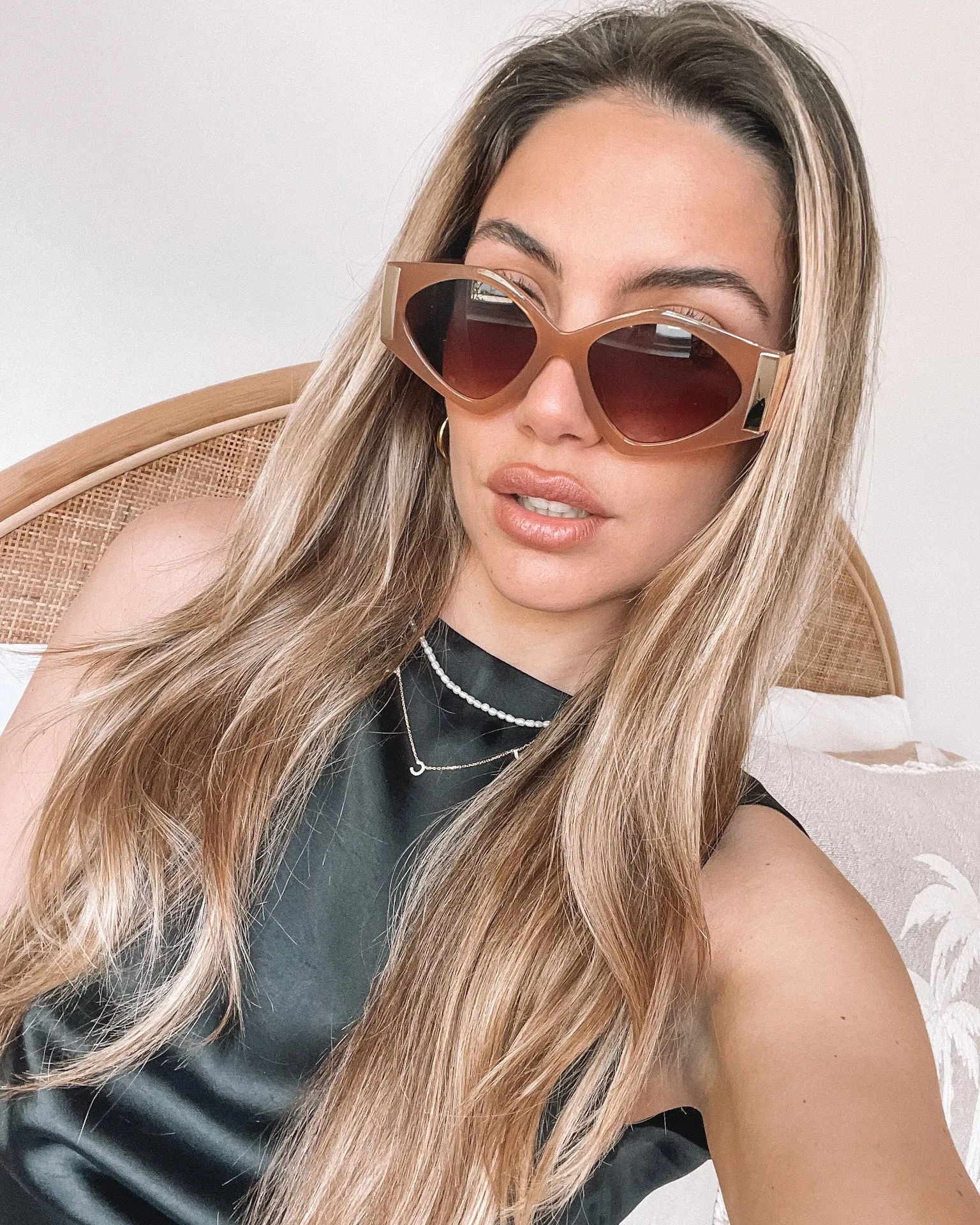 Brown cat-eye sunglasses with thick, glossy frames and dark brown tinted lenses, featuring gold accents on the corners, displayed on a light beige surface against a plain white background.