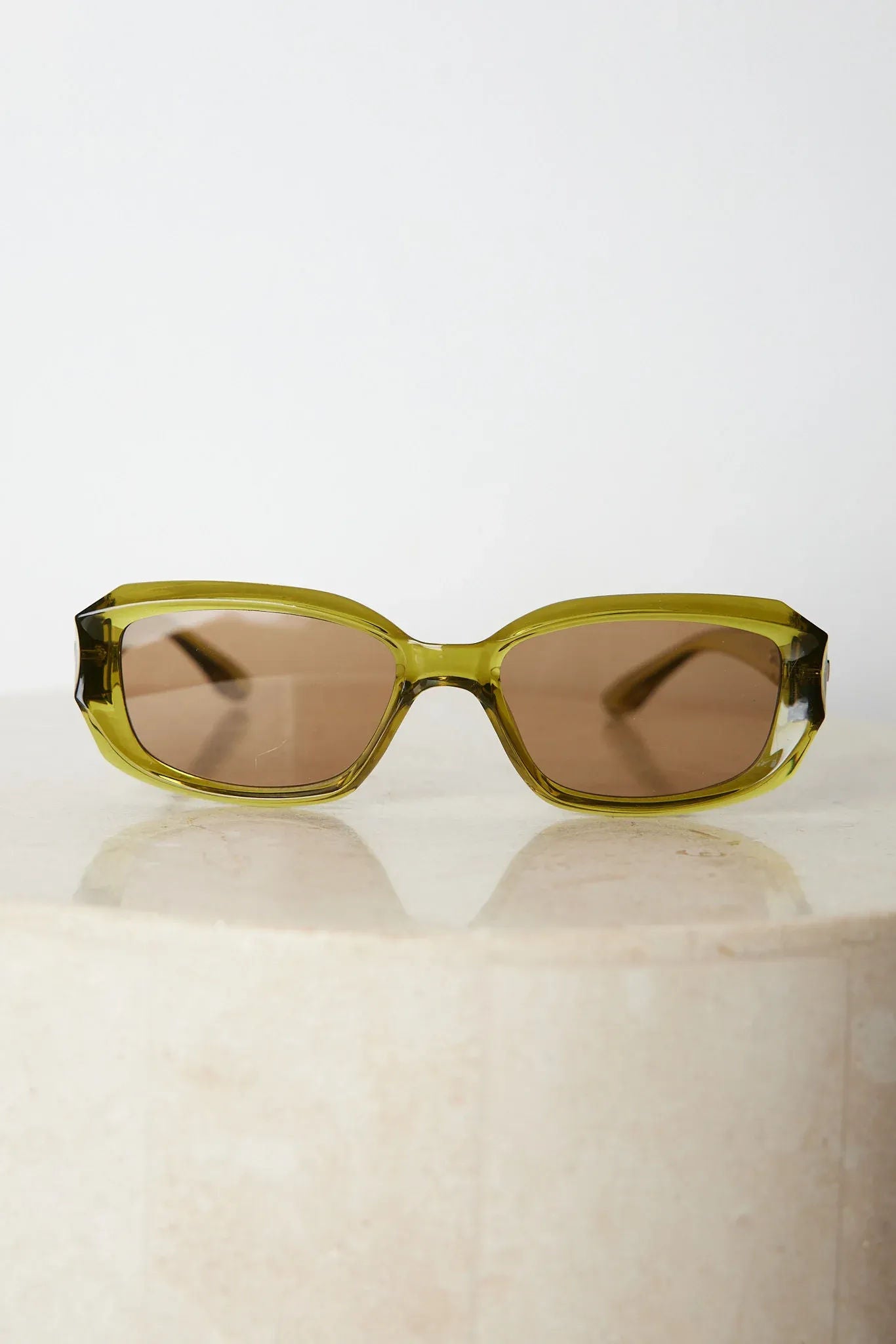 Rectangular sunglasses with translucent olive green frames and brown tinted lenses, displayed on a light beige stone surface against a plain white background.