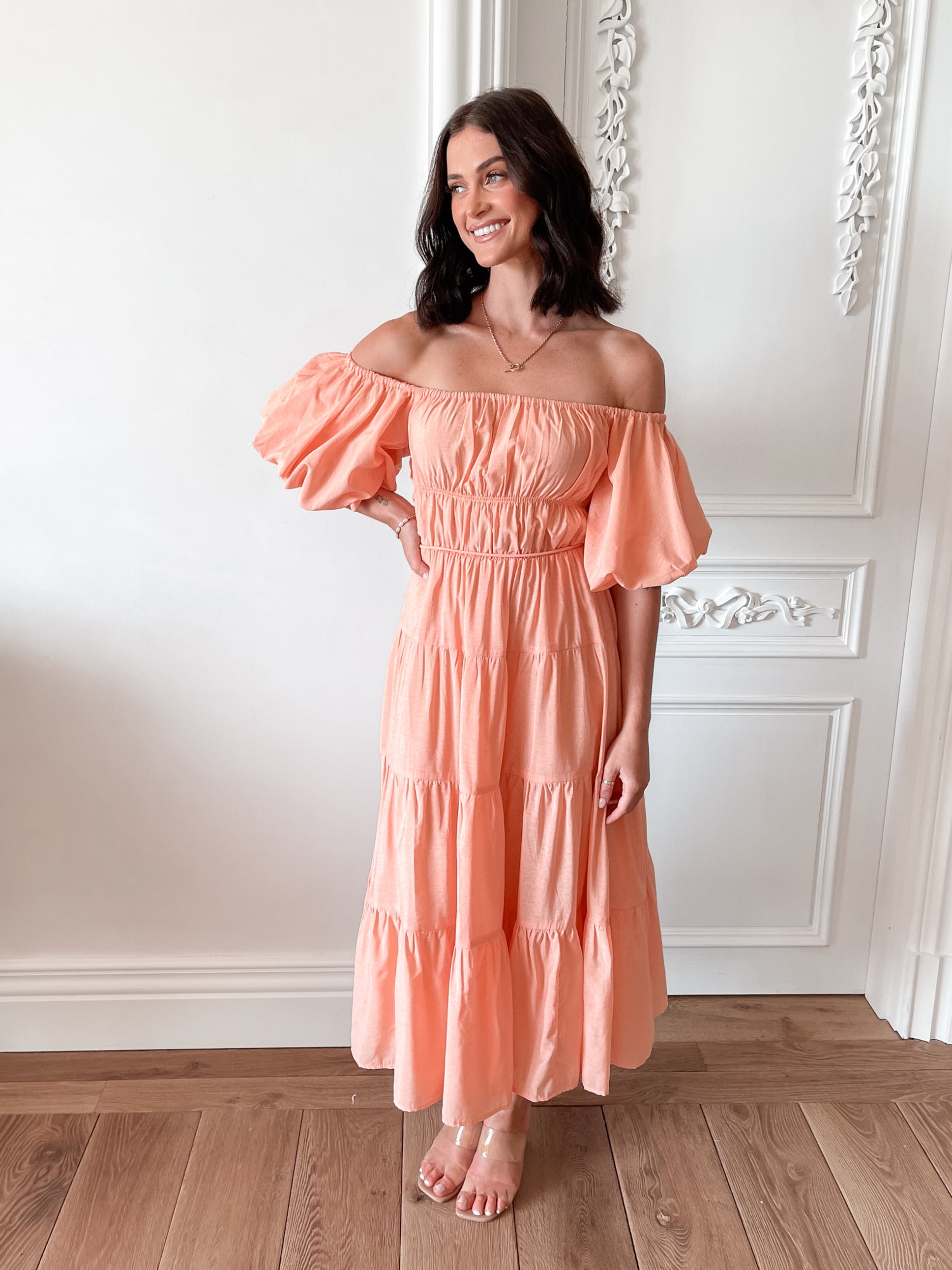 A woman with shoulder-length dark hair stands in a beautifully decorated room with white ornate wall paneling and wooden flooring, wearing an off-the-shoulder peach midi dress with puffed sleeves and a tiered skirt. She is smiling and gently holding one side of her dress, giving a soft and elegant pose.