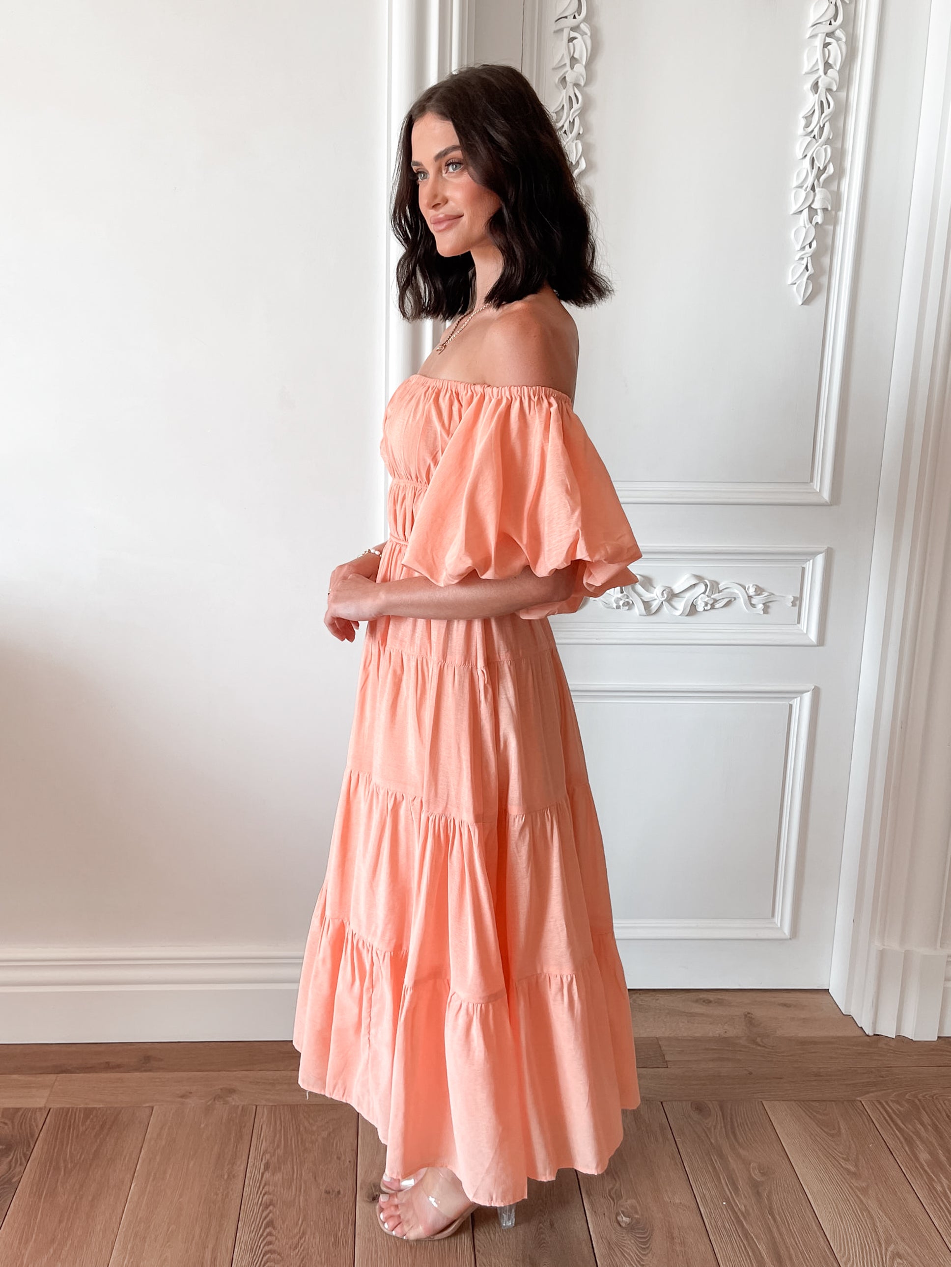 A woman with shoulder-length dark hair stands in a beautifully decorated room with white ornate wall paneling and wooden flooring, wearing an off-the-shoulder peach midi dress with puffed sleeves and a tiered skirt. She is smiling and gently holding one side of her dress, giving a soft and elegant pose.