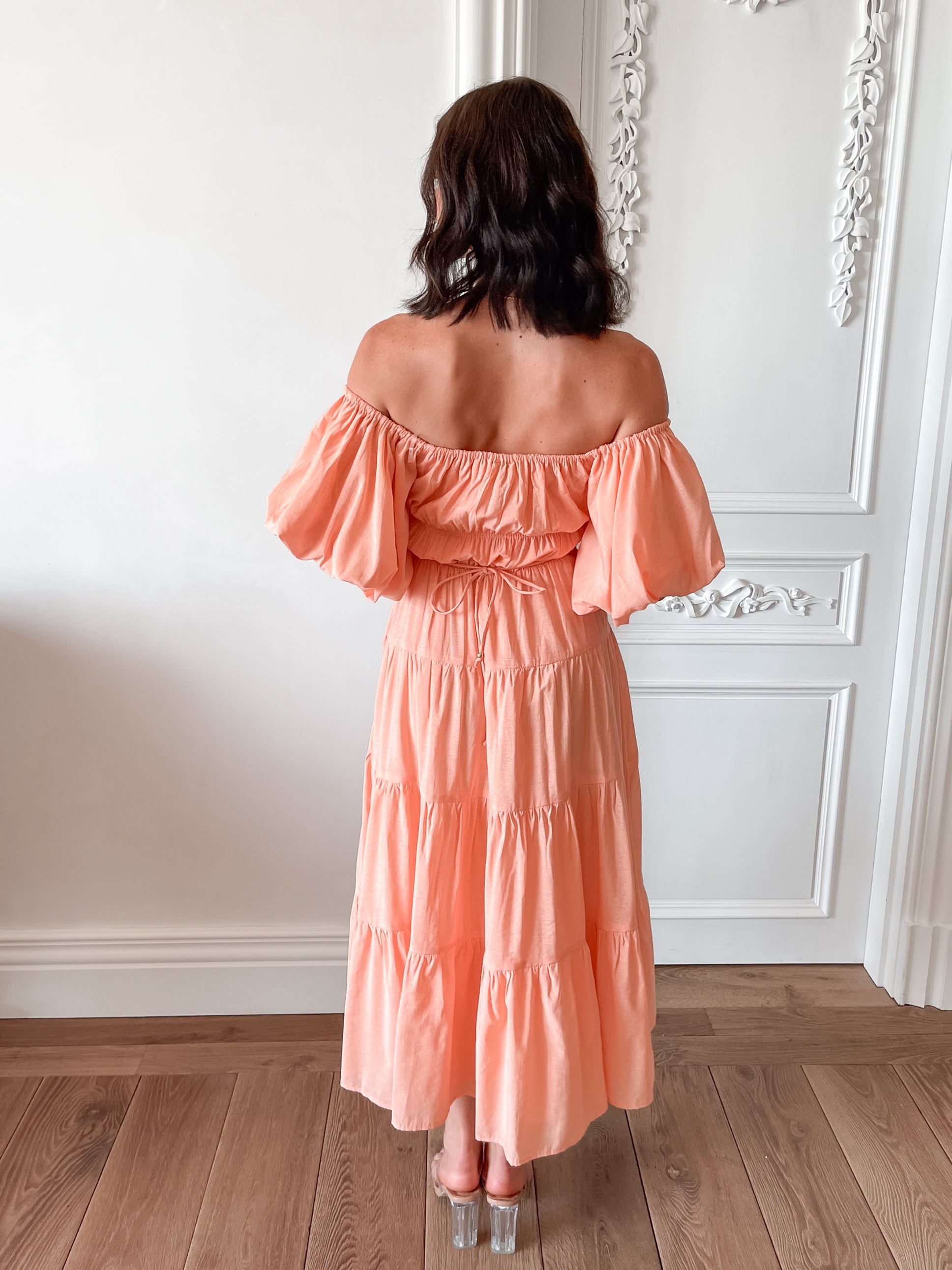 A woman with shoulder-length dark hair stands in a beautifully decorated room with white ornate wall paneling and wooden flooring, wearing an off-the-shoulder peach midi dress with puffed sleeves and a tiered skirt. She is smiling and gently holding one side of her dress, giving a soft and elegant pose.