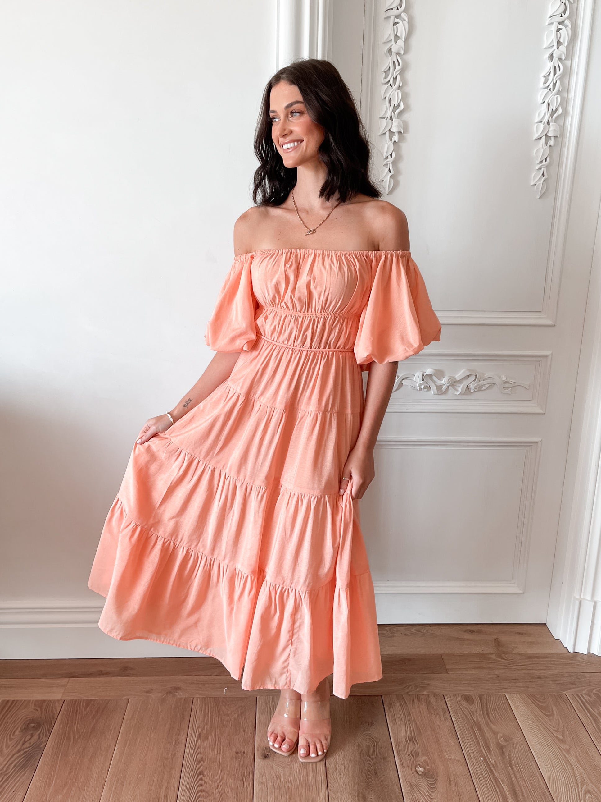 A woman with shoulder-length dark hair stands in a beautifully decorated room with white ornate wall paneling and wooden flooring, wearing an off-the-shoulder peach midi dress with puffed sleeves and a tiered skirt. She is smiling and gently holding one side of her dress, giving a soft and elegant pose.