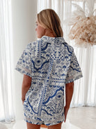 A woman with blonde, wavy hair stands in a bright, modern room with floor-to-ceiling sheer white curtains and neutral-toned decor. She is wearing a coordinated short-sleeve button-up shirt and matching shorts set featuring a blue and white floral and geometric print. She has a relaxed expression with one hand in her pocket, exuding a stylish and effortless vibe.