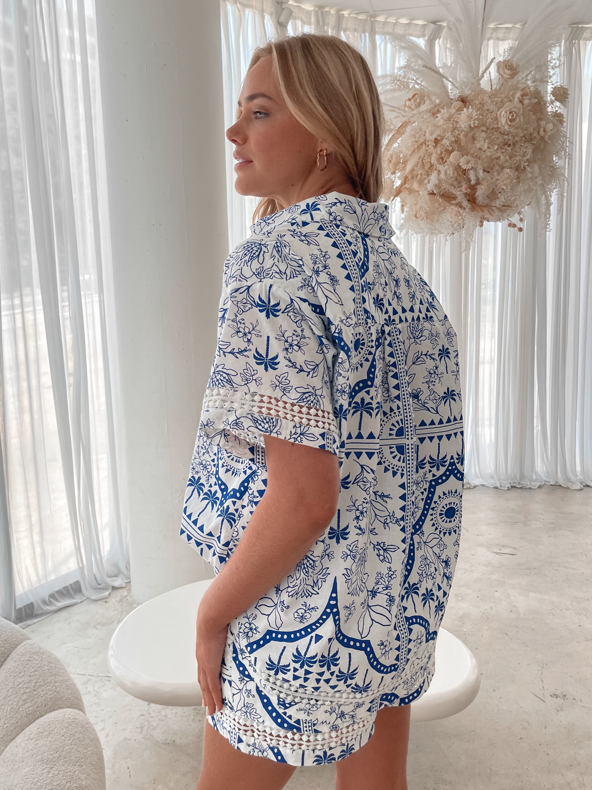 A woman with blonde, wavy hair stands in a bright, modern room with floor-to-ceiling sheer white curtains and neutral-toned decor. She is wearing a coordinated short-sleeve button-up shirt and matching shorts set featuring a blue and white floral and geometric print. She has a relaxed expression with one hand in her pocket, exuding a stylish and effortless vibe.