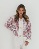 A woman with long, wavy brown hair stands against a plain white background, wearing a textured cardigan with a red, pink, and navy checkered pattern. The cardigan features large gold buttons and front pockets, layered over a white knit top. She pairs it with light cream-colored pants and poses with her hands in her pockets, gazing slightly to the side with a composed expression.