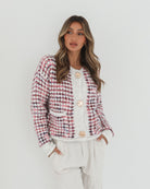 A woman with long, wavy brown hair stands against a plain white background, wearing a textured cardigan with a red, pink, and navy checkered pattern. The cardigan features large gold buttons and front pockets, layered over a white knit top. She pairs it with light cream-colored pants and poses with her hands in her pockets, gazing slightly to the side with a composed expression.