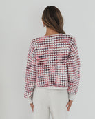 A woman with long, wavy brown hair stands against a plain white background, wearing a textured cardigan with a red, pink, and navy checkered pattern. The cardigan features large gold buttons and front pockets, layered over a white knit top. She pairs it with light cream-colored pants and poses with her hands in her pockets, gazing slightly to the side with a composed expression.