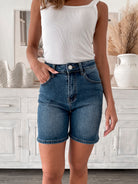 A smiling woman with long, wavy hair stands indoors in front of a white distressed wooden sideboard decorated with a large shell sculpture, a white vase holding dried palm leaves, and a round wall clock. She is wearing a white sleeveless tank top paired with high-waisted denim shorts and clear strap heels, with one hand resting in her pocket. The setting has a cozy, neutral-toned decor with a woven basket to the side and a patterned rug underfoot.
