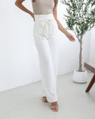 A woman wearing high-waisted, wide-leg white trousers with a paper bag waist and a fabric tie belt, paired with a sleeveless beige ribbed top and nude heels, standing in a minimalistic room with a potted tree and a wooden bench.