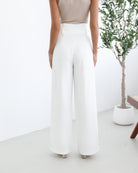 A woman wearing high-waisted, wide-leg white trousers with a paper bag waist and a fabric tie belt, paired with a sleeveless beige ribbed top and nude heels, standing in a minimalistic room with a potted tree and a wooden bench.