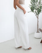 A woman wearing high-waisted, wide-leg white trousers with a paper bag waist and a fabric tie belt, paired with a sleeveless beige ribbed top and nude heels, standing in a minimalistic room with a potted tree and a wooden bench.