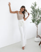 A woman wearing high-waisted, wide-leg white trousers with a paper bag waist and a fabric tie belt, paired with a sleeveless beige ribbed top and nude heels, standing in a minimalistic room with a potted tree and a wooden bench.