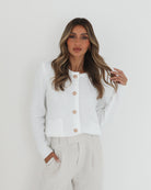 A woman wearing a white textured knit cardigan with front buttons and two small front pockets, paired with high-waisted light beige trousers. She stands against a plain white background, with one hand in her pocket and the other touching her hair.