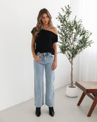 Woman wearing light-wash flared jeans and black heeled ankle boots, paired with a sleeveless black top, standing indoors near a potted plant and a wooden chair.