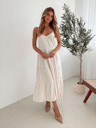 A woman wearing a beige, midi-length Sonya dress with a V neckline, adjustable spaghetti straps, a shirred back, and a tiered skirt. The dress is sleeveless and the photo shows the dress in a relaxed fit style.