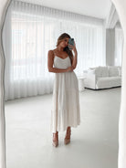 A woman wearing a beige, midi-length Sonya dress with a V neckline, adjustable spaghetti straps, a shirred back, and a tiered skirt. The dress is sleeveless and the photo shows the dress in a relaxed fit style.