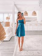 A woman standing in a minimalist setting wearing a teal mini dress with slim straps and a sweetheart neckline.