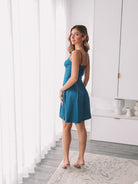 A woman standing in a minimalist setting wearing a teal mini dress with slim straps and a sweetheart neckline.