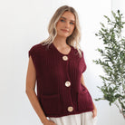 A woman wearing a sleeveless wine-colored knit top with round neckline and center-front buttons, paired with light-colored pants.