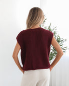 A woman wearing a sleeveless wine-colored knit top with round neckline and center-front buttons, paired with light-colored pants.