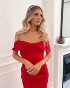 Elegant woman wearing a red off-shoulder gown, perfect for formal events, weddings, evening balls, and special occasions, standing in a sophisticated room with wooden flooring and a fireplace.