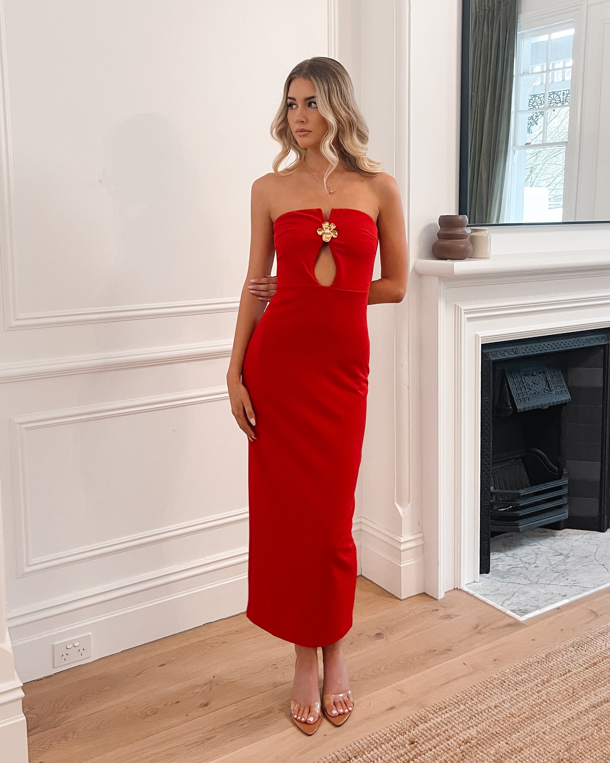 Elegant woman in a red strapless evening gown, perfect for weddings, formal events, balls, galas, and special occasions, standing in a sophisticated room with wooden flooring and a fireplace.