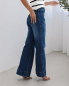 A woman standing in a room with light decor, wearing a striped top and mid-blue Yzabel Jeans with a high waist fit and wide leg silhouette.