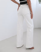 A female model wearing white high-waisted jeans with brown contrasting stitches, paired with a striped black and white top.