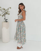 A woman standing in a room with a white background, wearing a mint floral dress with bow straps and a square neckline.