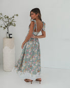 A woman standing in a room with a white background, wearing a mint floral dress with bow straps and a square neckline.