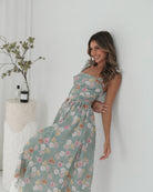 A woman standing in a room with a white background, wearing a mint floral dress with bow straps and a square neckline.