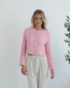 A woman wearing a pink Valdivia Jacket with long sleeves, round neckline, and center-front buttons. The jacket has a crop length and she is paired with light-colored trousers.