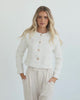 A woman sitting in a chair wearing a white Valdivia Jacket with long sleeves, round neckline, and center-front buttons. The jacket has a crop length and she is paired with a light-colored pants.