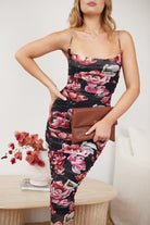 A woman wearing a strapless, form-fitting midi dress with a bold floral print in shades of pink and red on a black background. She is standing beside a white sofa and a light wood coffee table with a vase of red flowers, in a cozy room with neutral-toned decor and a woven rug.