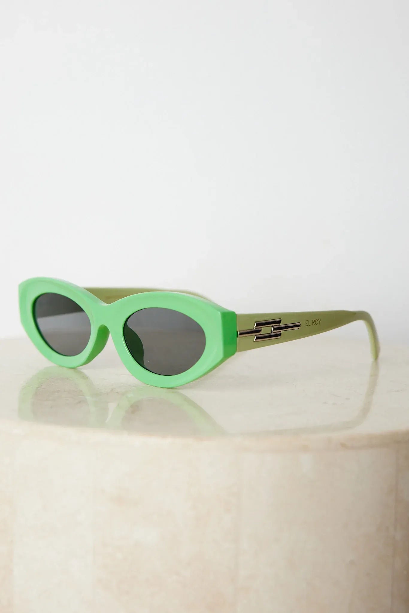 Close-up of a pair of bright green cat-eye sunglasses with dark lenses, placed on a smooth beige surface with a plain white background.